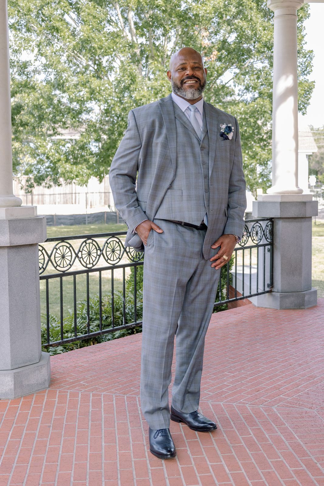 Handsome Black groom in gray plaid three-piece suit with boutonniere posing outdoors at wedding venue.