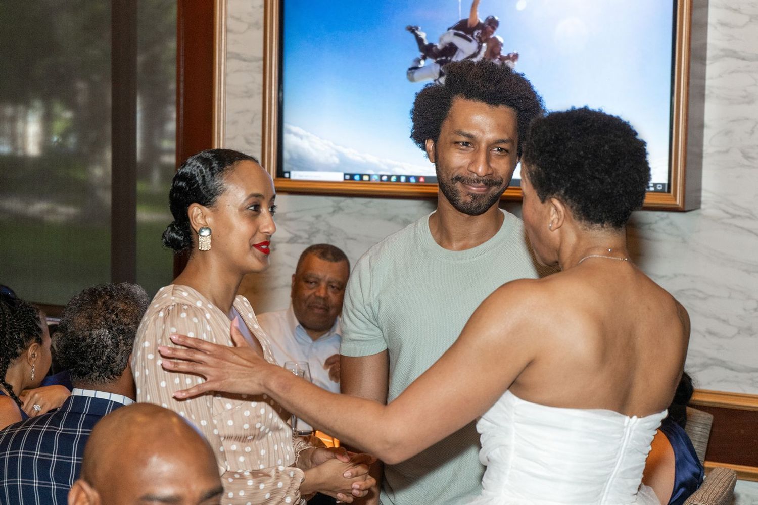 Guests in formal attire greet bride in white dress at wedding reception with TV screen in background.