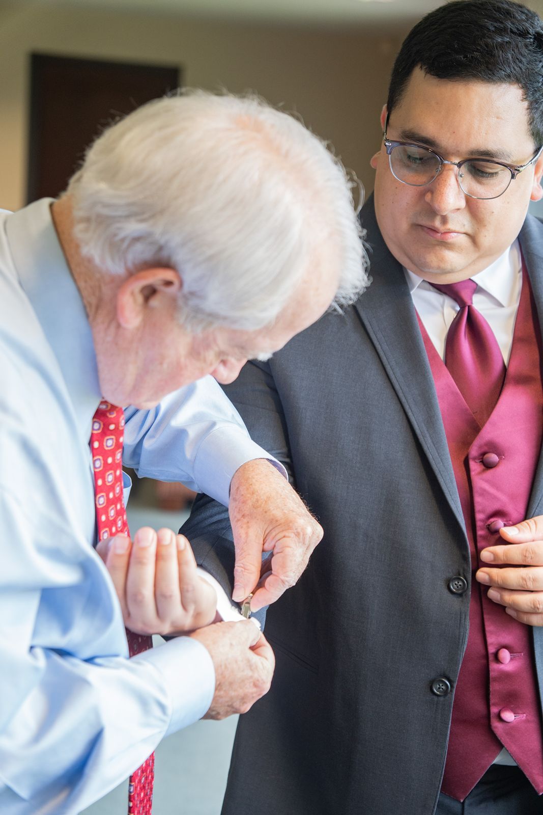 Two men in formal attire stand together as one adjusts the other's cufflinks or sleeve buttons.