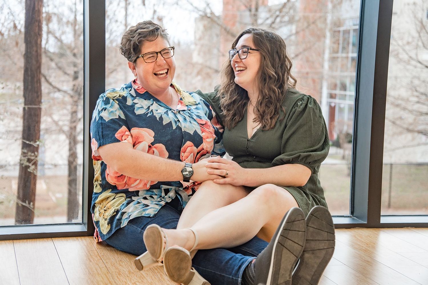 A couple laughs together while sitting near large windows in a bright indoor space.