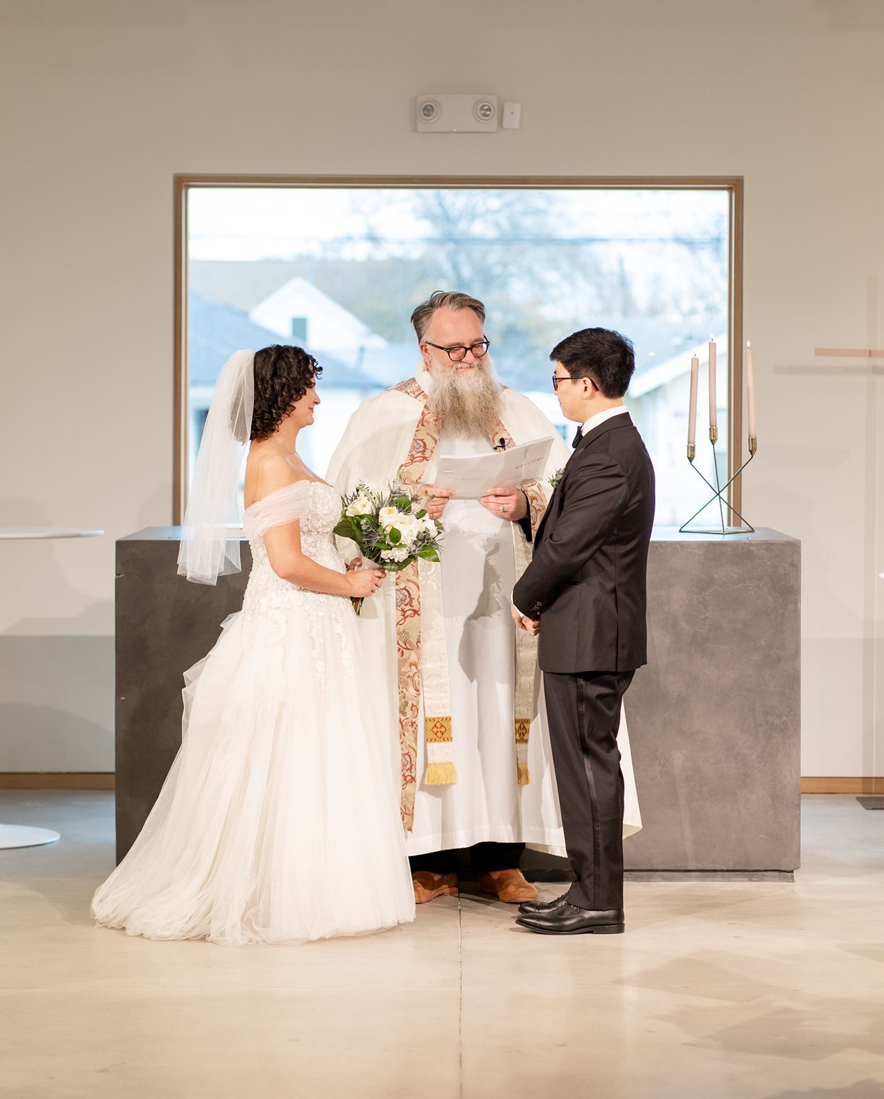 A wedding ceremony unfolds in a modern interior space with gray concrete walls and large windows overlooking snowy mountains.