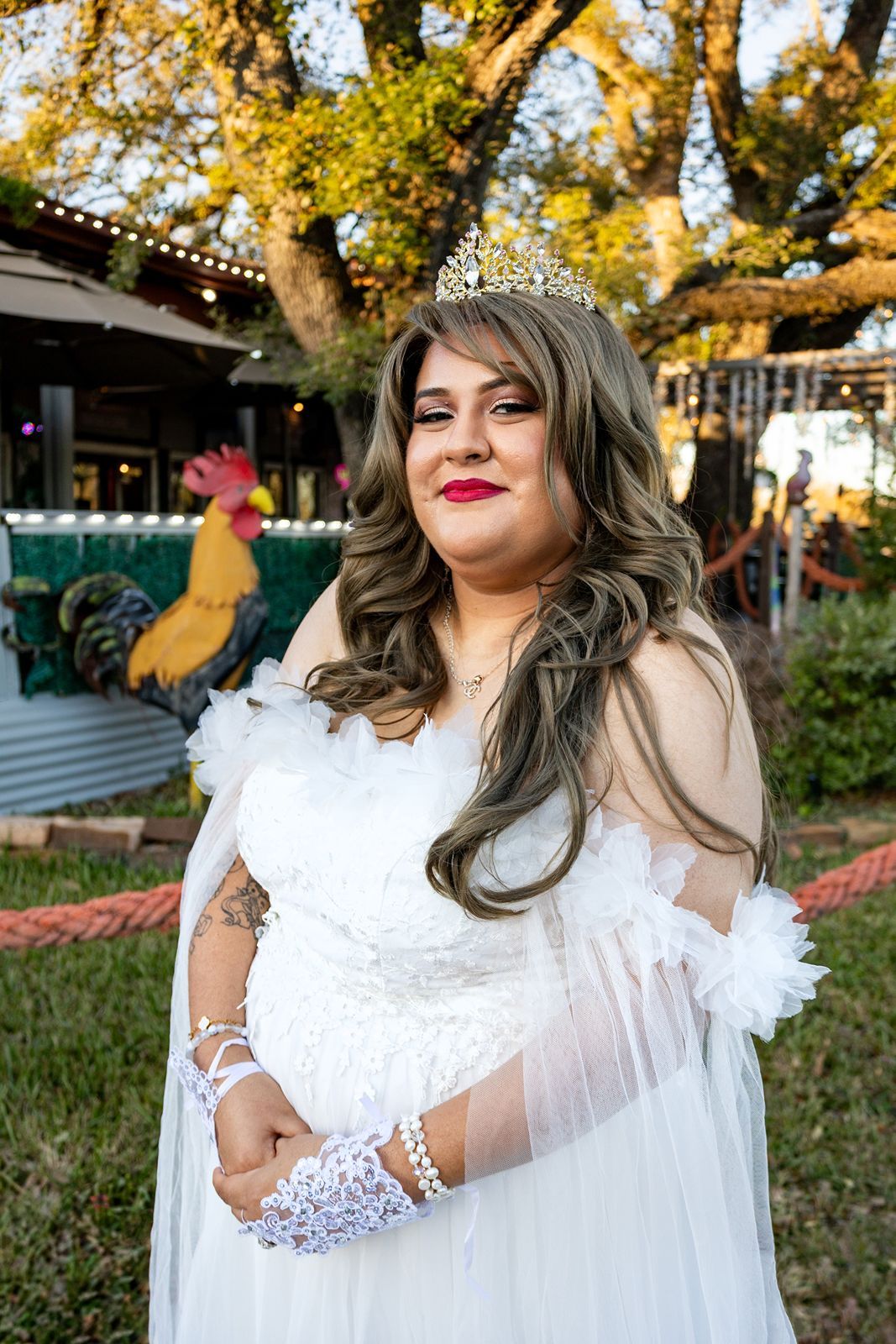 Woman in white wedding dress and tiara poses outdoors near rooster statue and string lights.