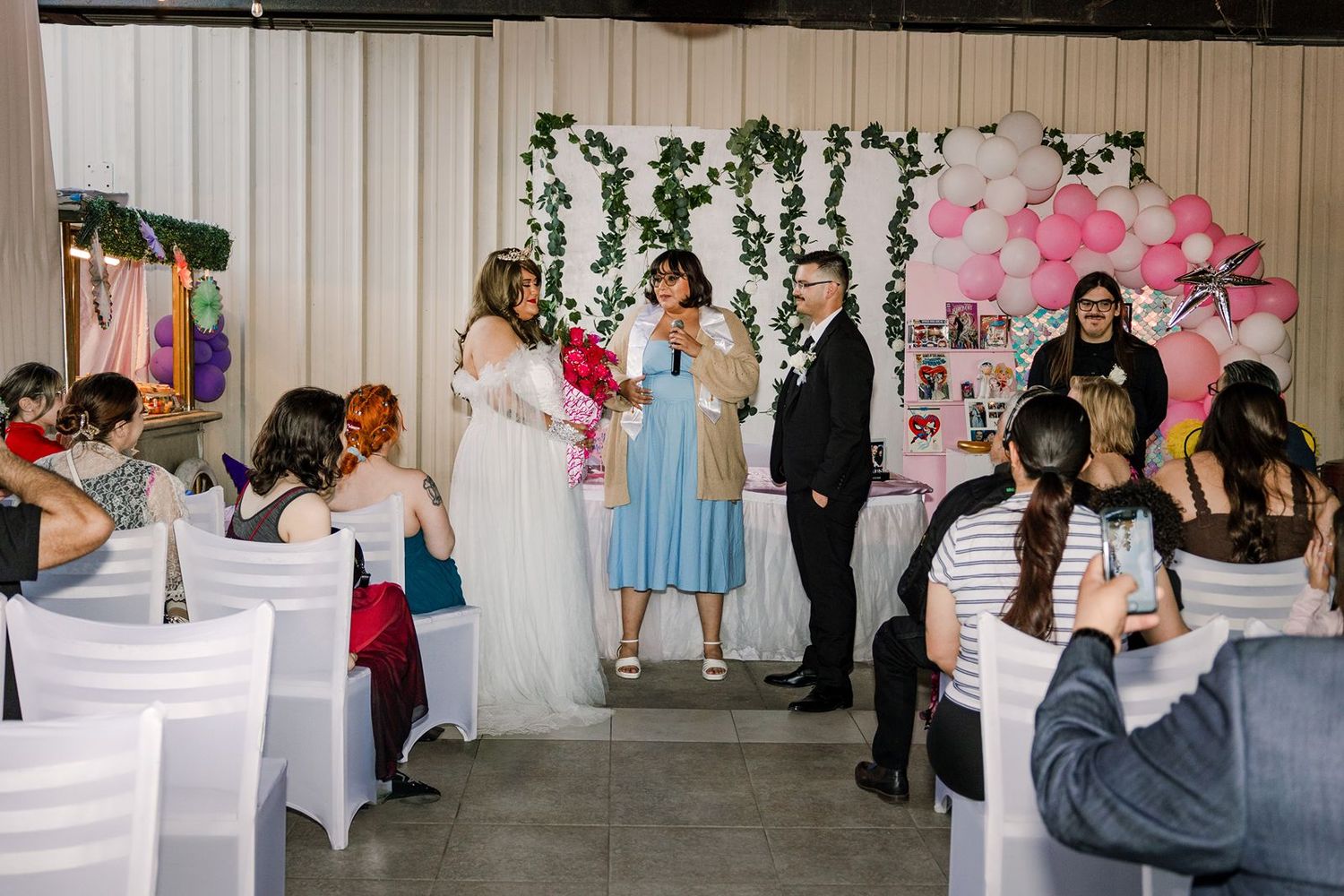 Bride and groom exchange vows at indoor wedding ceremony with pink balloon arch and floral backdrop.
