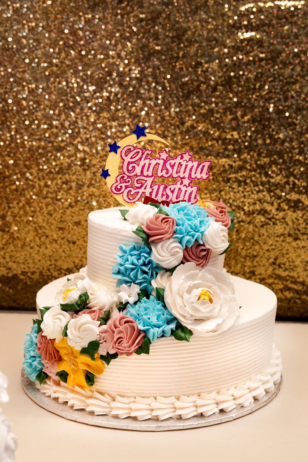 Two-tier white wedding cake with colorful floral decorations and Christina & Austin topper on gold background.