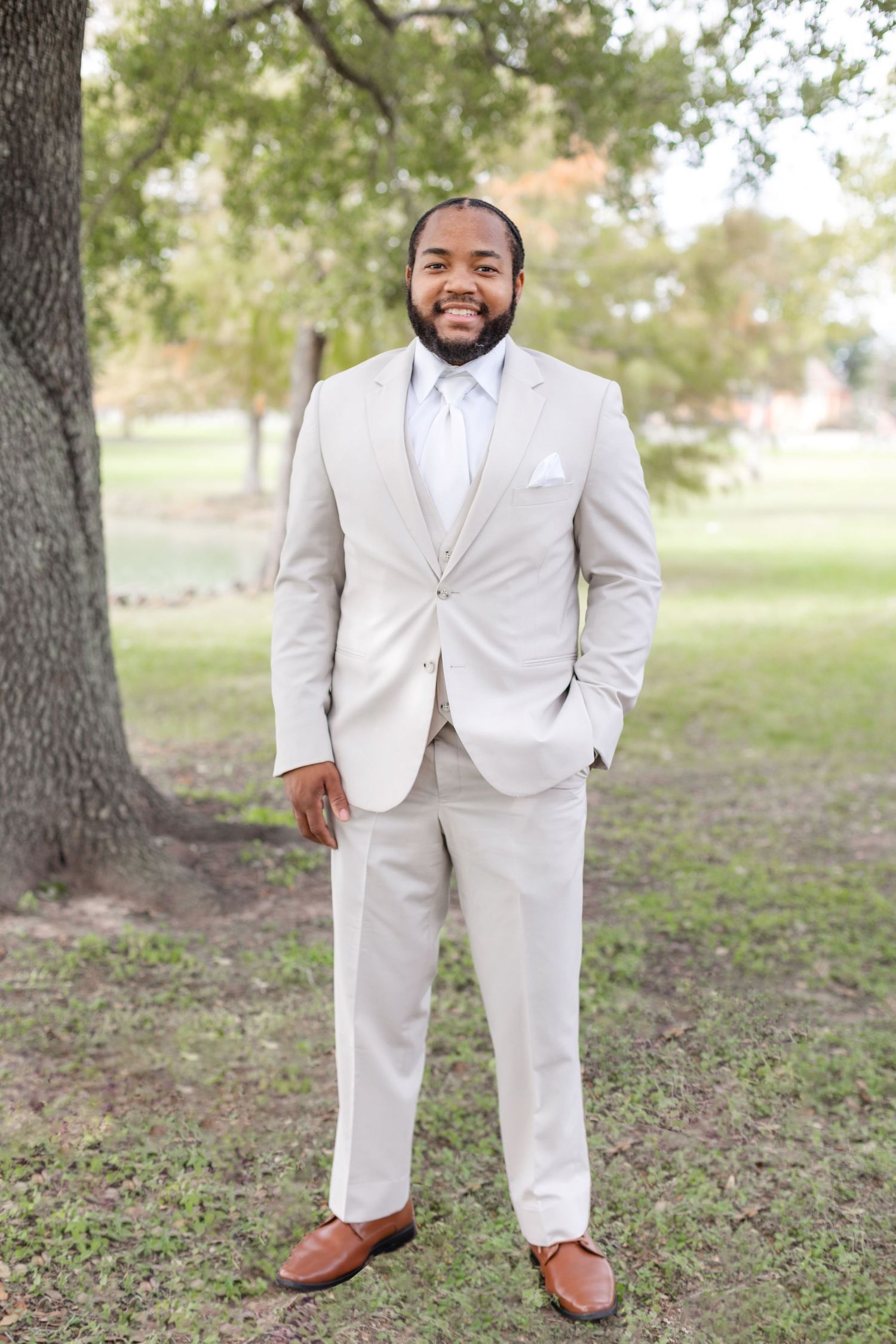 A groom wearing a classic white suit and brown shoes stands on leaf-covered ground under oak trees.