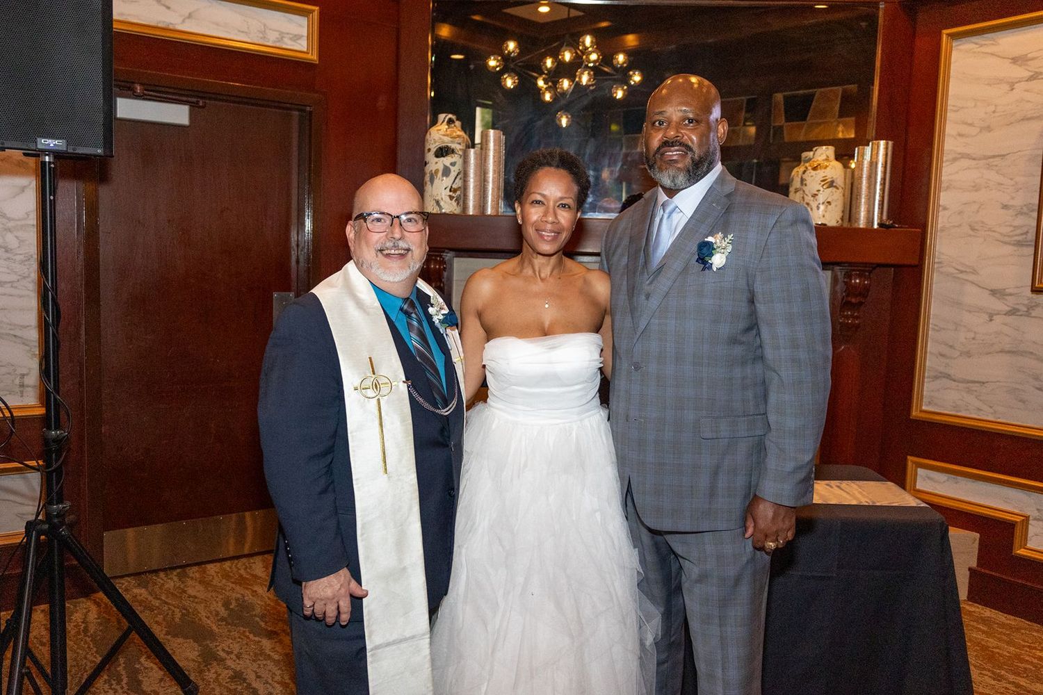 Bride in white gown poses with groom in gray suit and officiant wearing stole at indoor wedding reception.