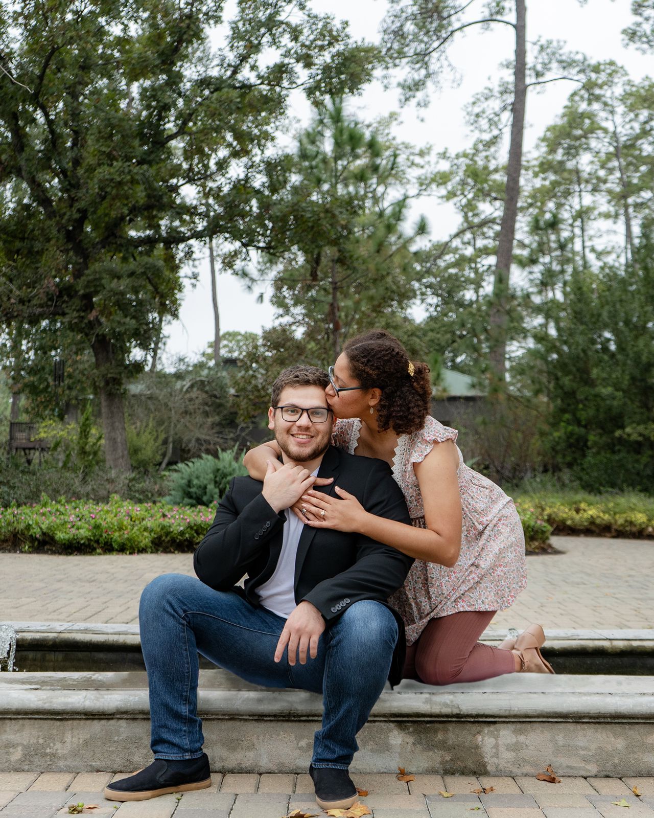 A couple shares a tender kiss on stone steps surrounded by lush greenery during an outdoor engagement photo session.