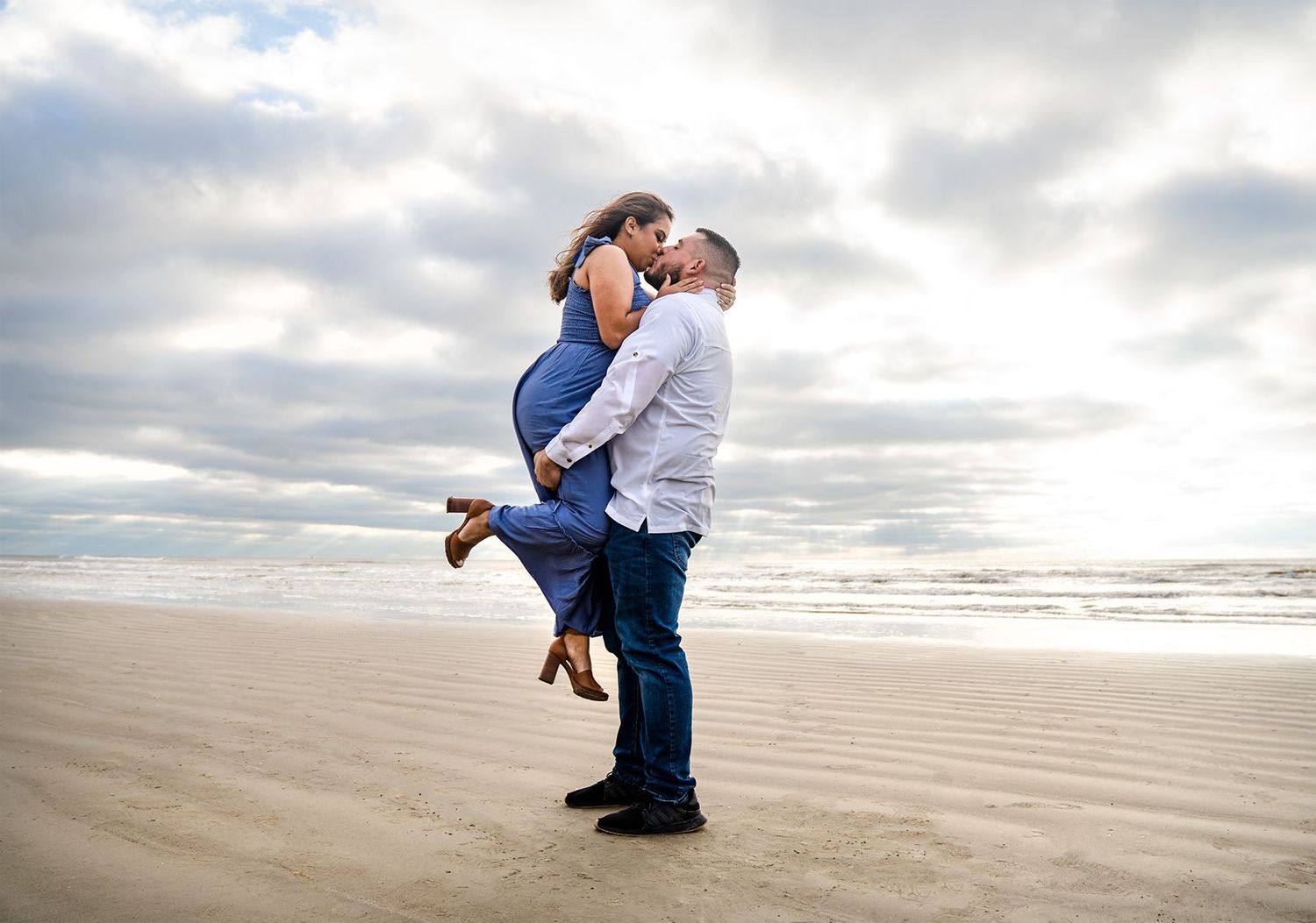 A couple embraces on a sandy beach at sunset with cloudy skies overhead.