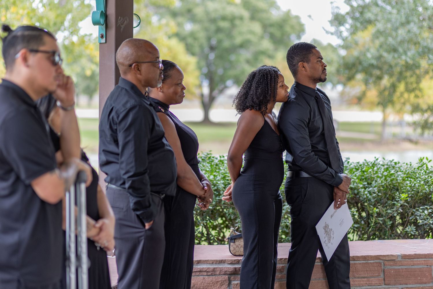 Wedding party members in black formal attire stand in line outdoors during a ceremony.
