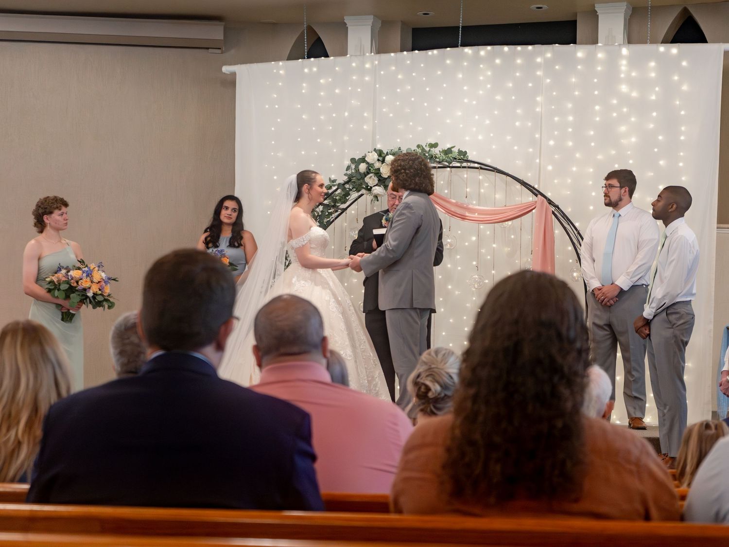 Wedding ceremony in progress at church altar with floral arch and light backdrop.
