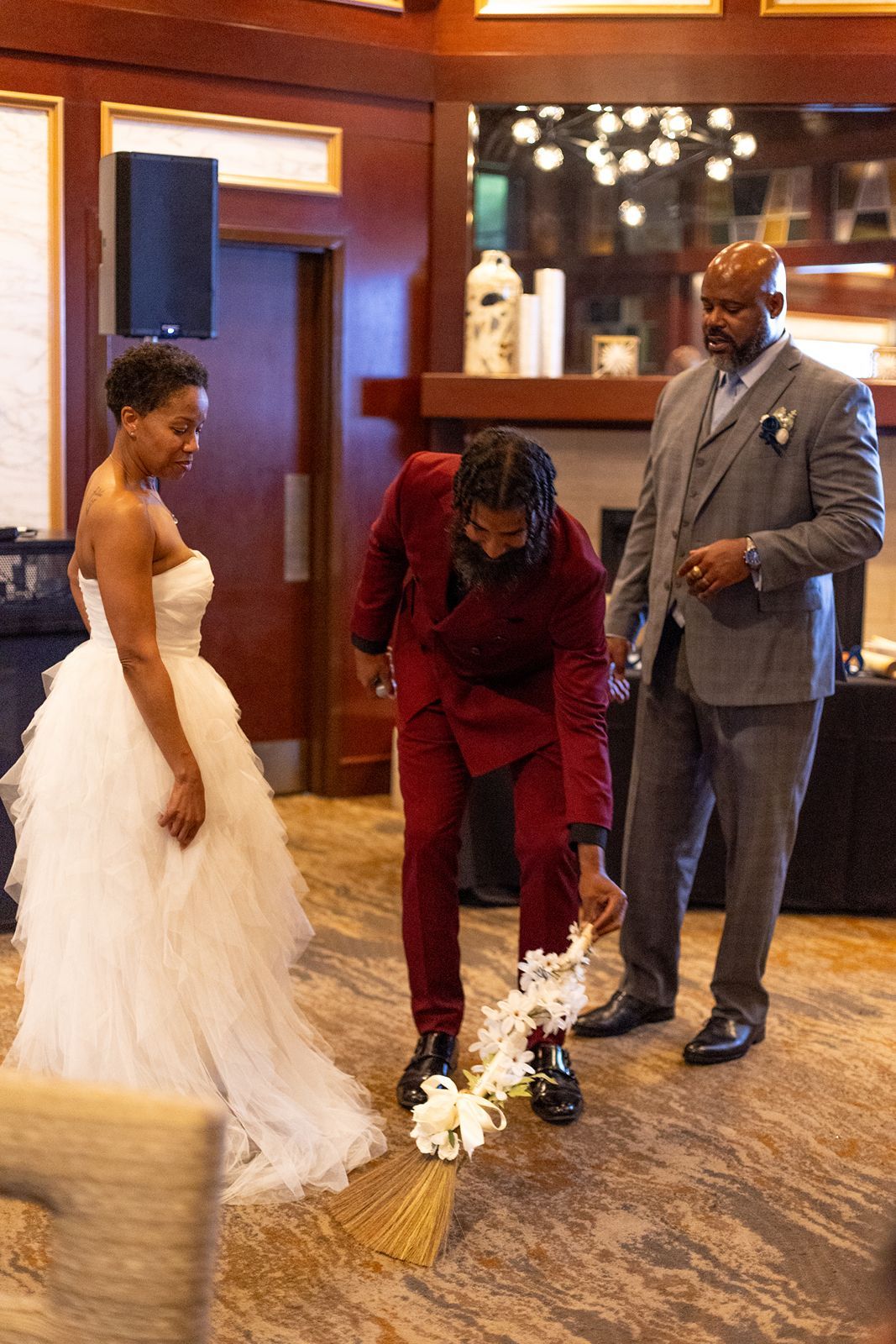 Wedding broom jumping ceremony with bride in white tulle gown, groom in red suit, and officiant in gray.