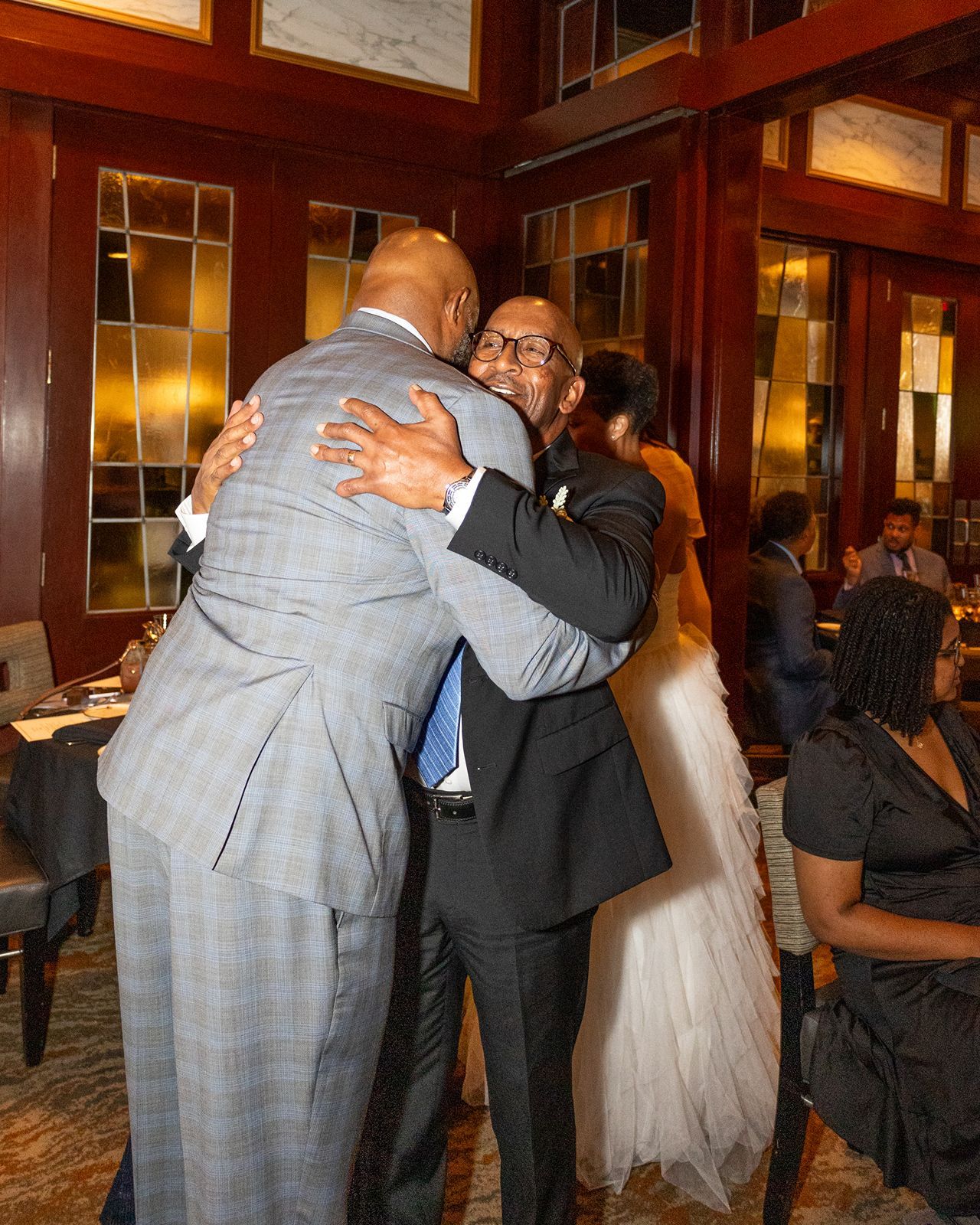 Two men share a warm embrace at an elegant wedding reception venue with wood paneling.