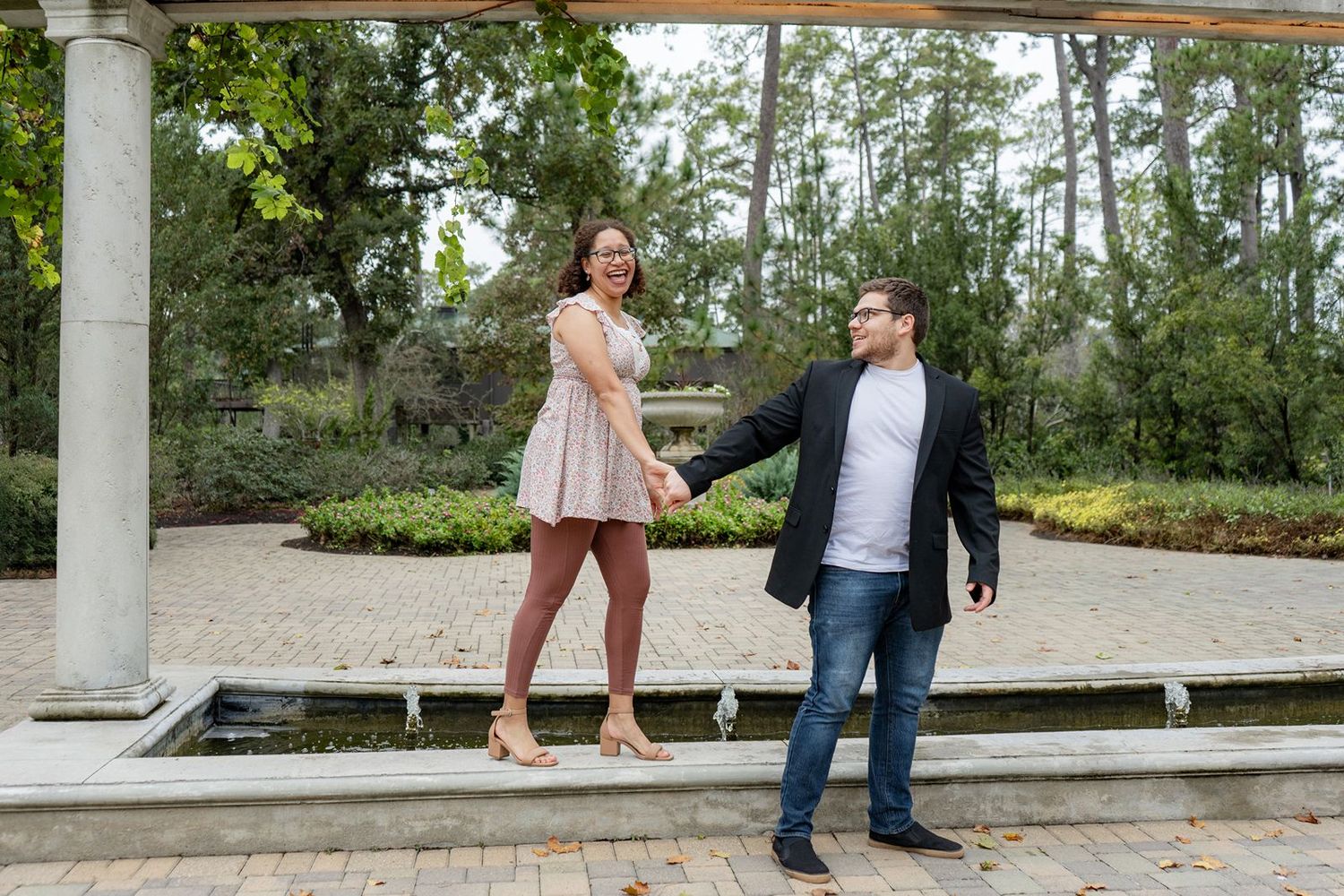 Couple holding hands and smiling in a lush garden gazebo setting, woman in pink dress, man in blazer and jeans.