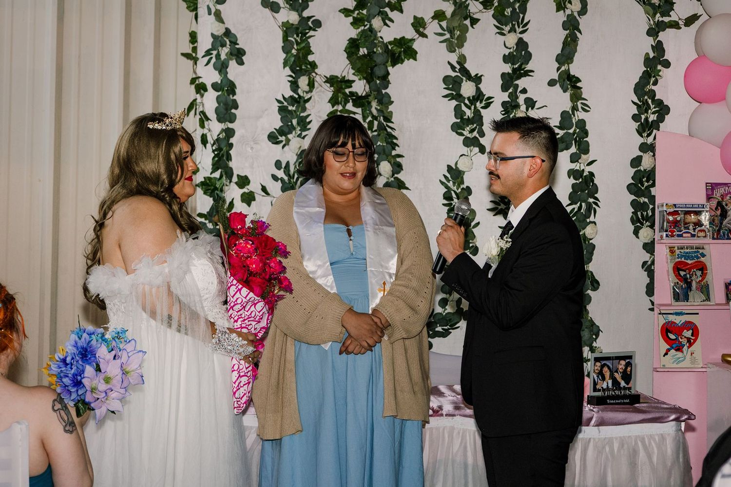Couple exchanges wedding vows before officiant in blue dress, with greenery backdrop and floral bouquets.
