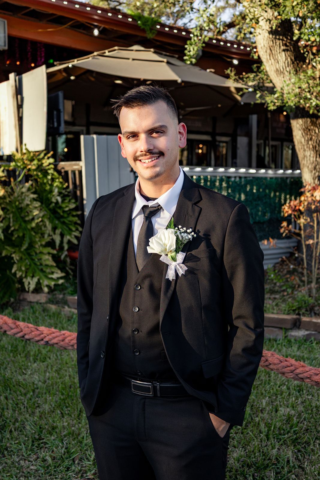 Young man in black suit with white boutonniere smiling outdoors at a wedding ceremony.