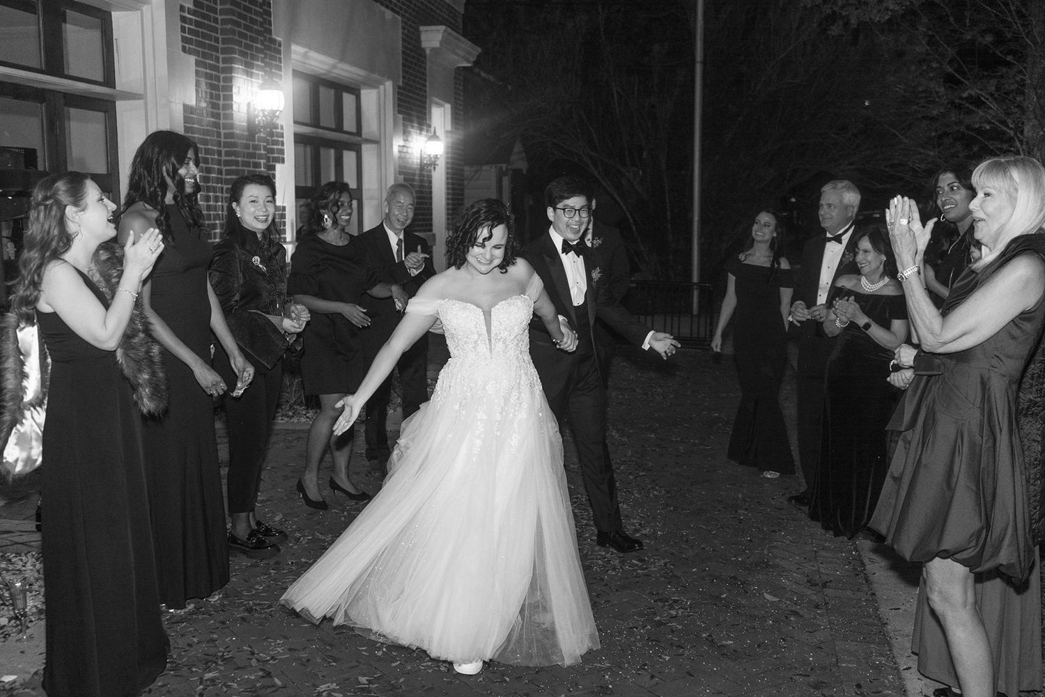 A couple dances together at a wedding reception while guests in dark dresses look on during an evening celebration.