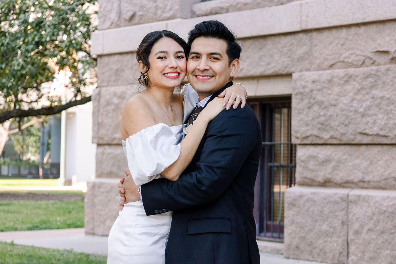 A joyful couple embraces outdoors in formal wedding attire in front of a stone building on a sunny day.