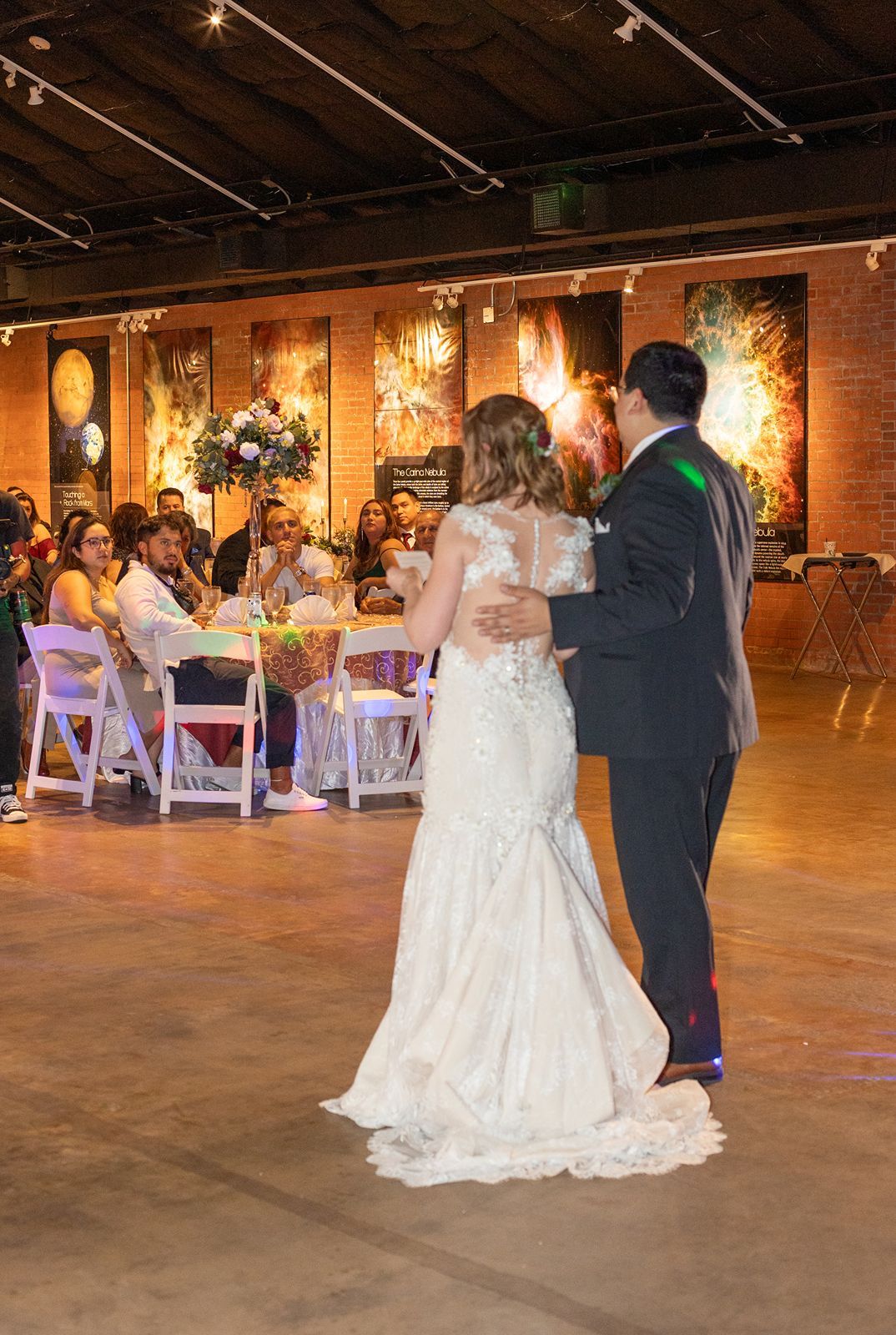 A wedding couple shares their first dance on a rustic industrial dance floor with string lights overhead and guests seated nearby.