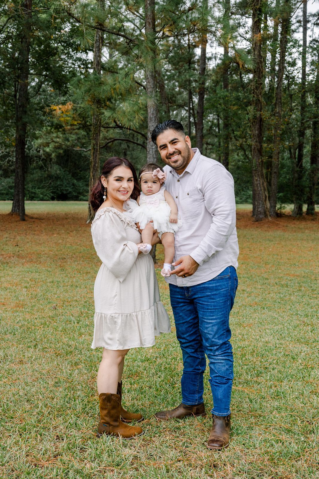 A family poses together outdoors in a wooded setting, wearing coordinated white and neutral outfits.