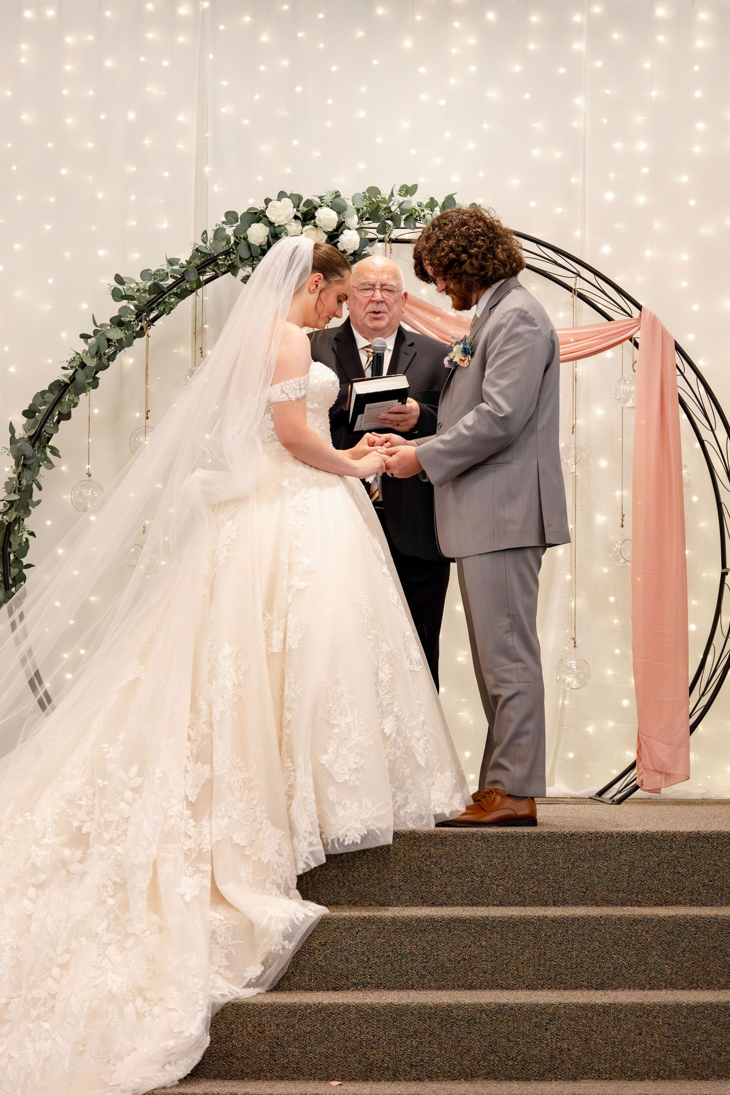 An intimate wedding ceremony moment captured beneath an arch adorned with greenery and fairy lights.