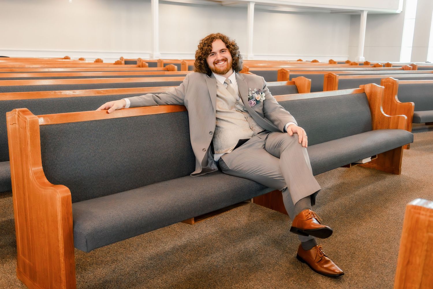 A relaxed posed photo on a church pew wearing a tailored grey suit.