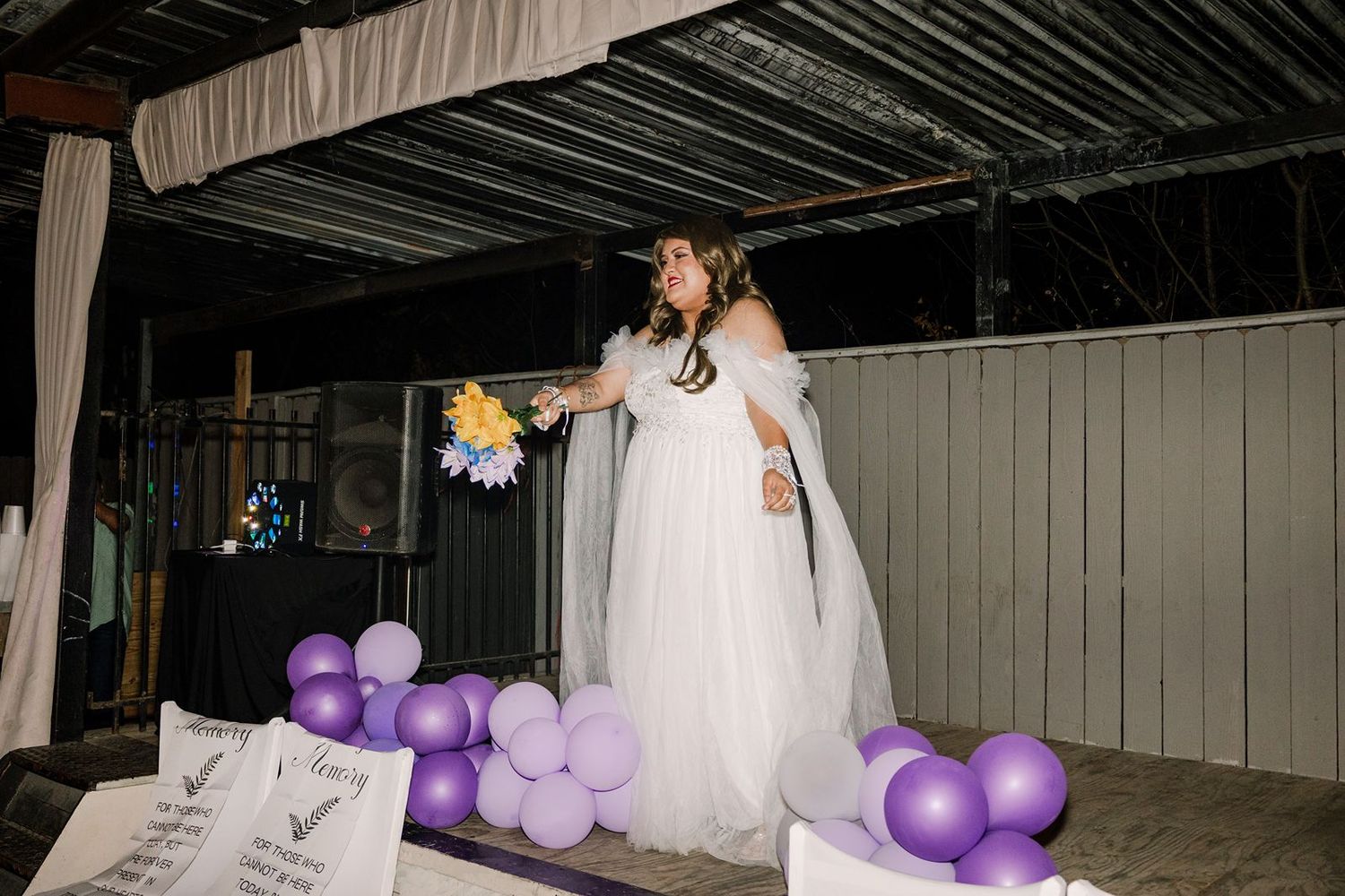 Bride in white gown tossing bouquet surrounded by purple balloons at outdoor wedding reception.