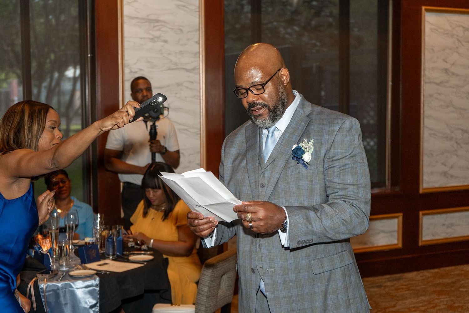 Man in gray suit reads from book at wedding reception while woman holds microphone nearby.