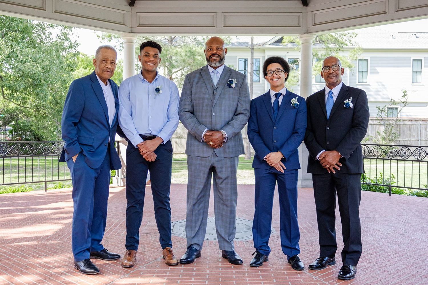 Five men in blue suits pose together under a gazebo at an outdoor wedding celebration.