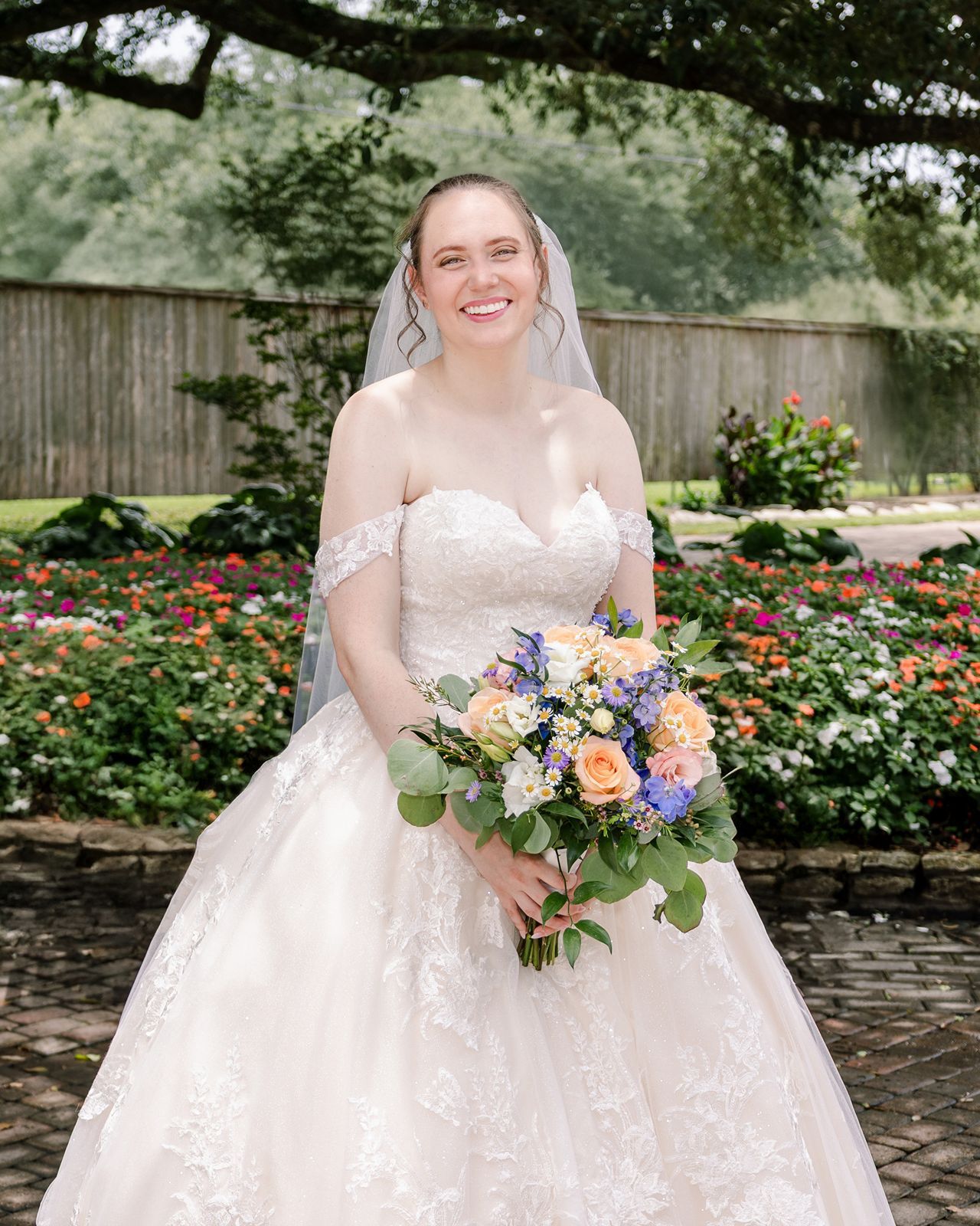 A bride in a lace wedding dress poses in a garden with colorful roses and stone walls, holding a vibrant bouquet.