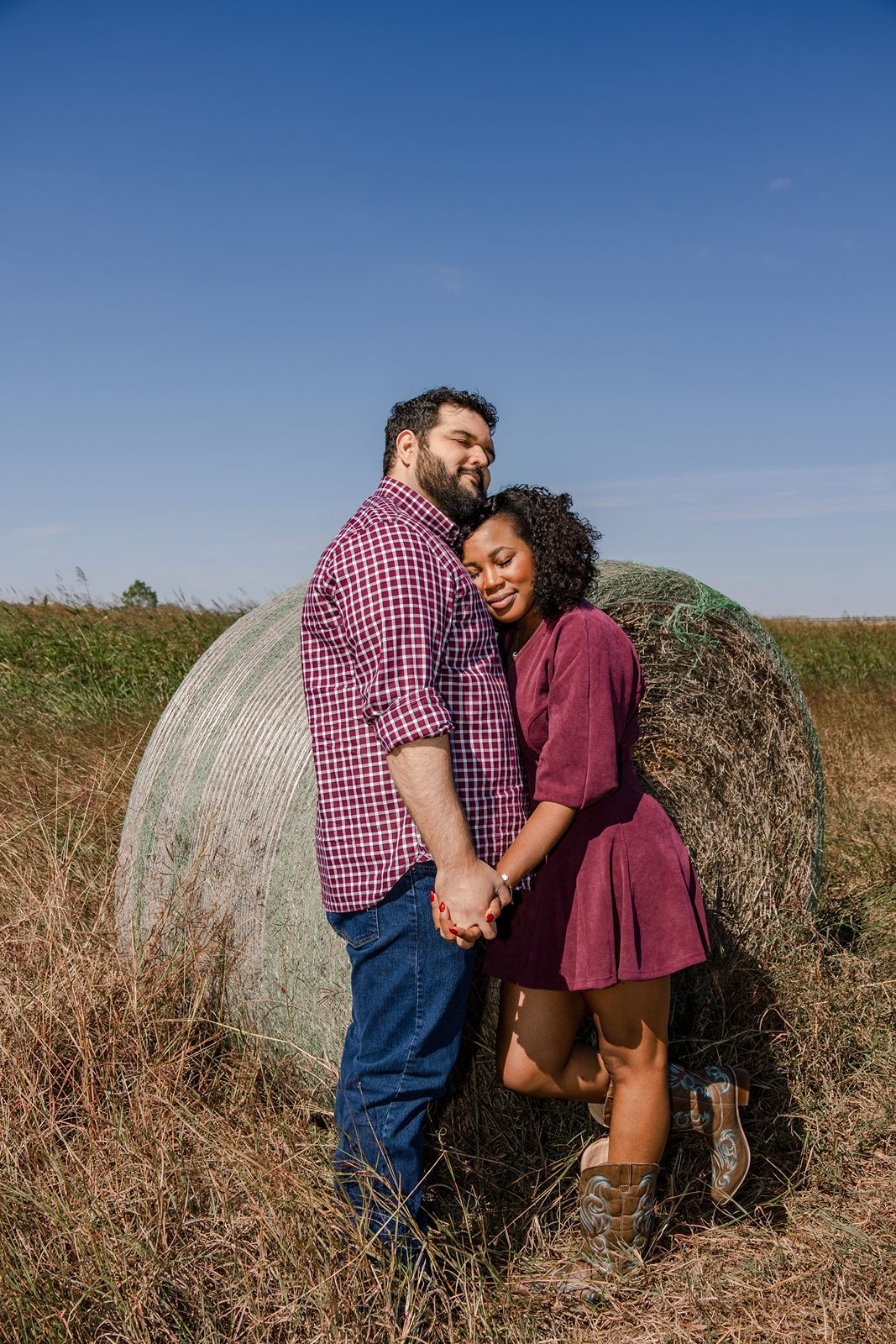A couple shares an intimate moment together while standing next to a large hay bale in a rural field.