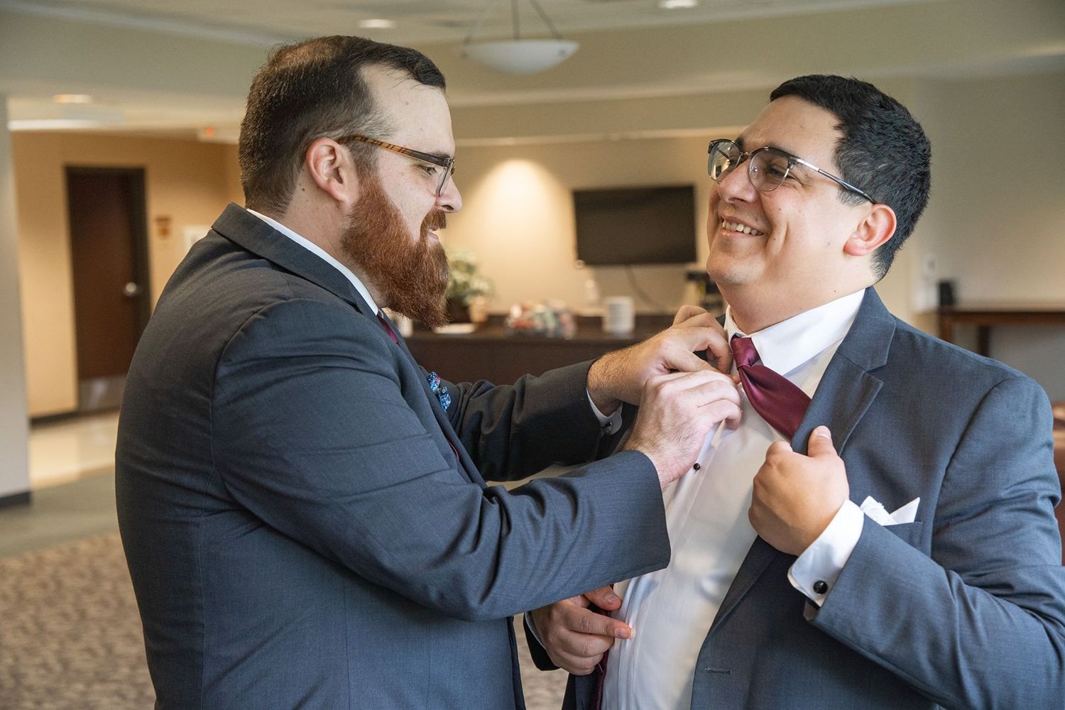 Two men in suits help each other adjust their neckties while sharing a lighthearted moment before an event.