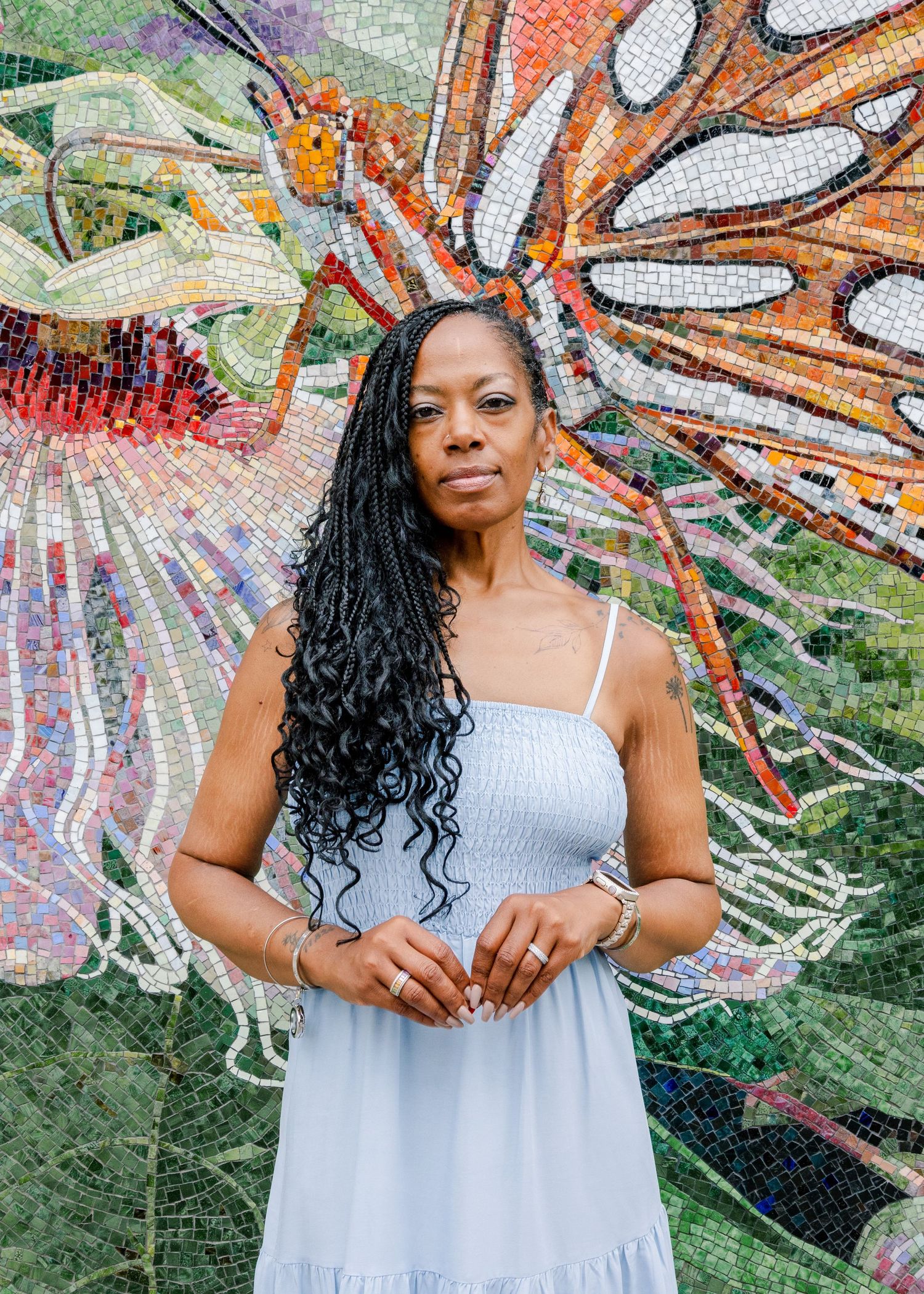Woman in light blue sundress poses against colorful mosaic wall background in garden setting.