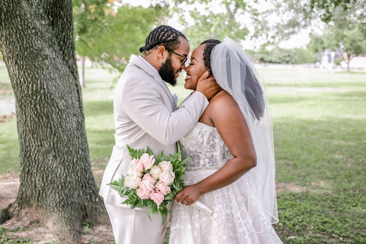 A romantic outdoor wedding moment under a tree with a bride holding a pink bouquet in a lace dress.