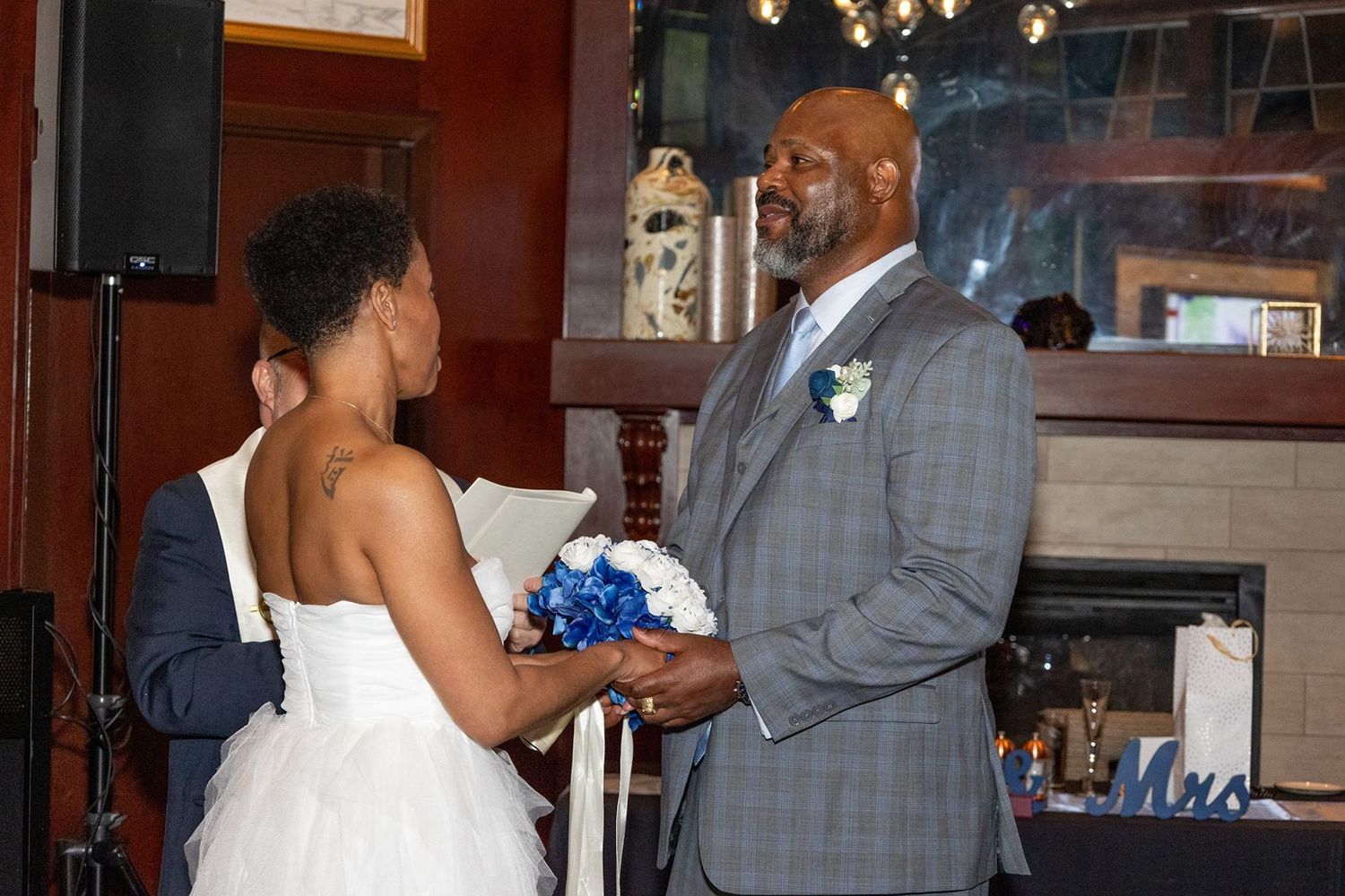Bride and groom exchanging vows indoors, groom in gray suit with blue boutonniere, bride holding blue and white bouquet.
