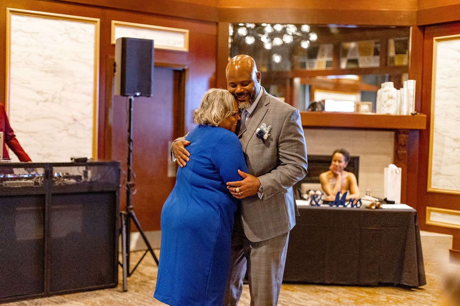 A man in a gray suit shares a warm embrace with a woman in a blue dress at a wedding reception.