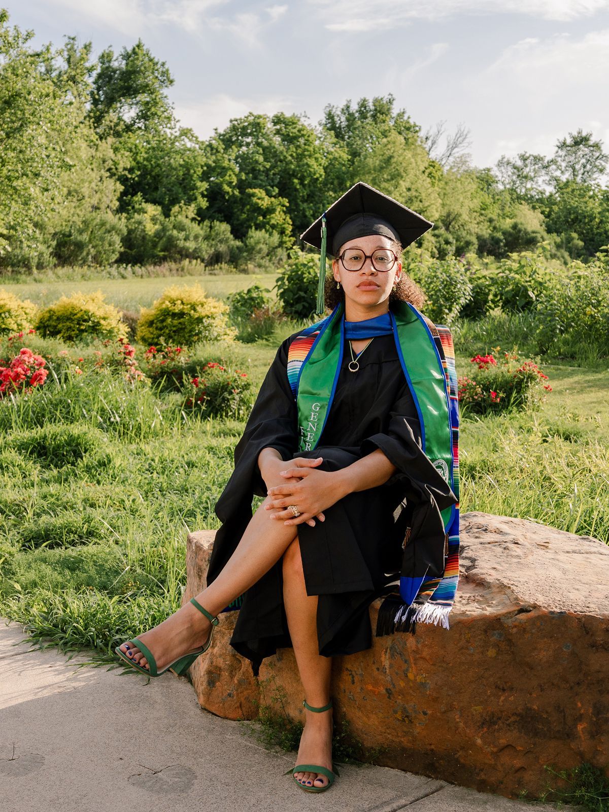Graduate in black cap and gown with colorful stoles sits on a rock in a lush green garden.
