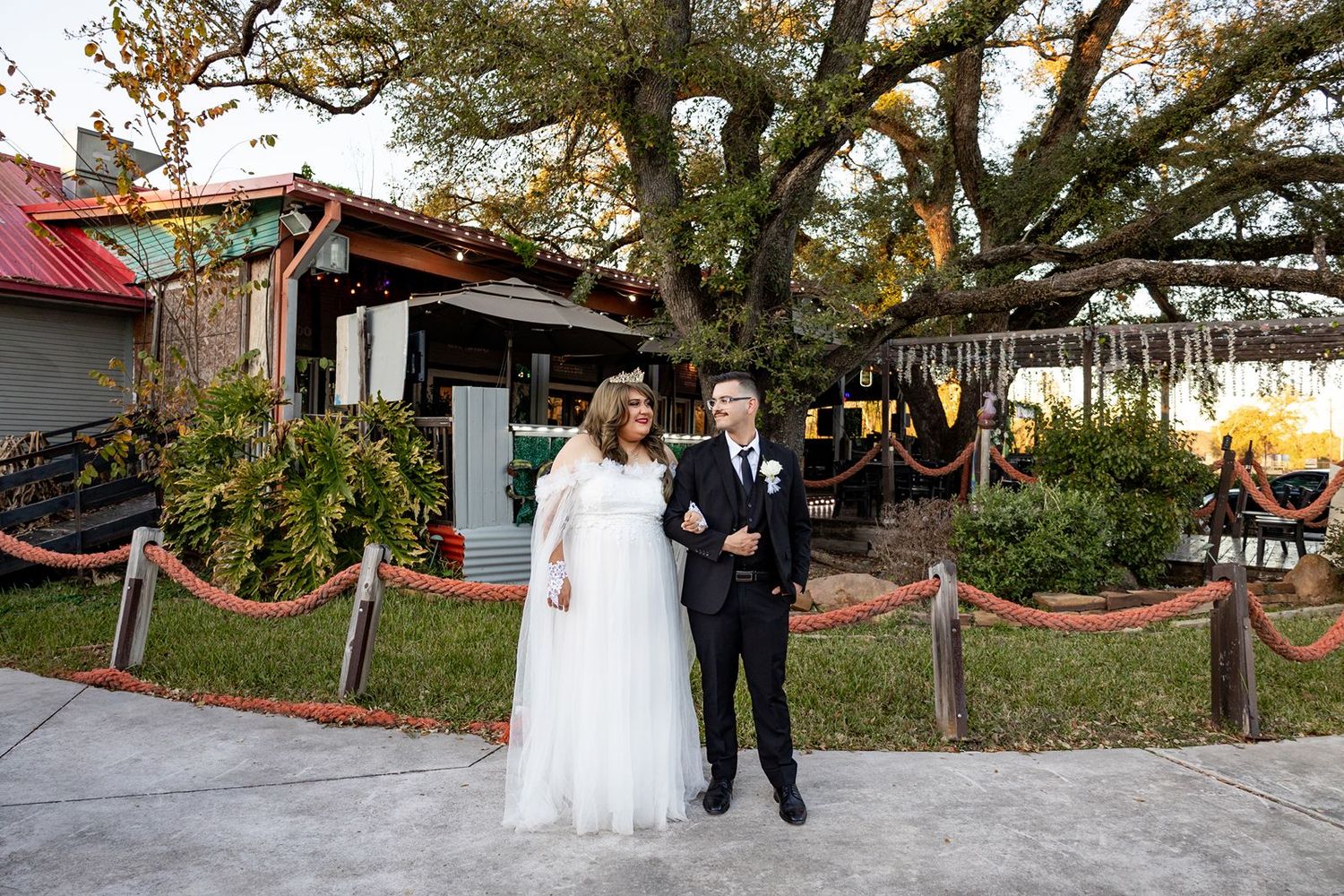 Bride in white gown and groom in black suit stand smiling outdoors near rustic venue with large oak tree.