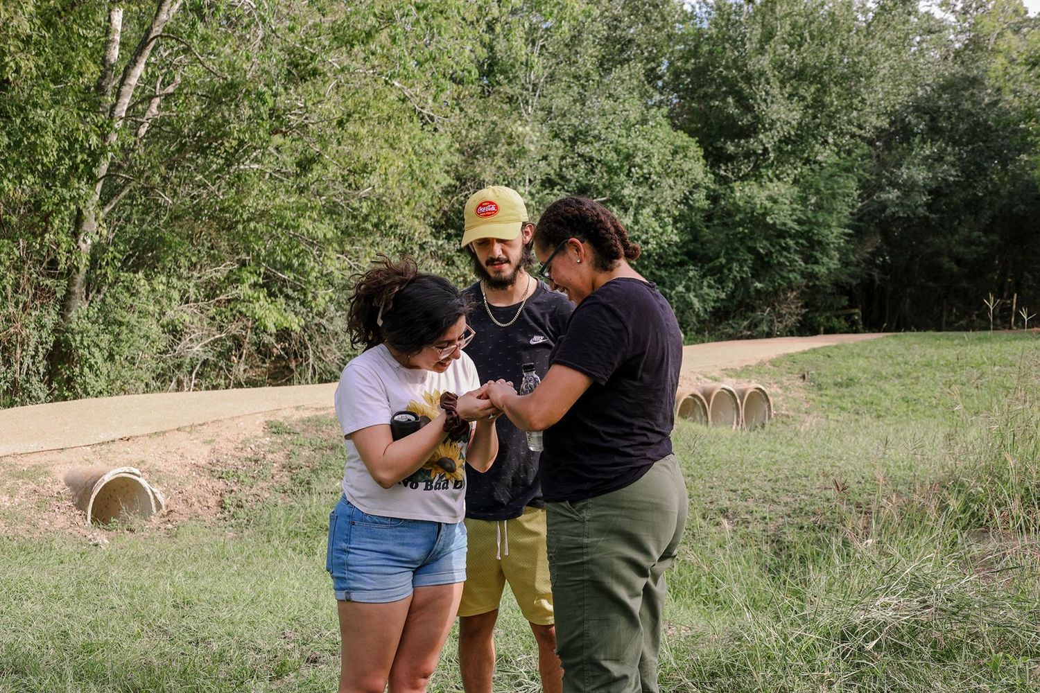 Three friends gathered outdoors examining an object together near a lush green forest.