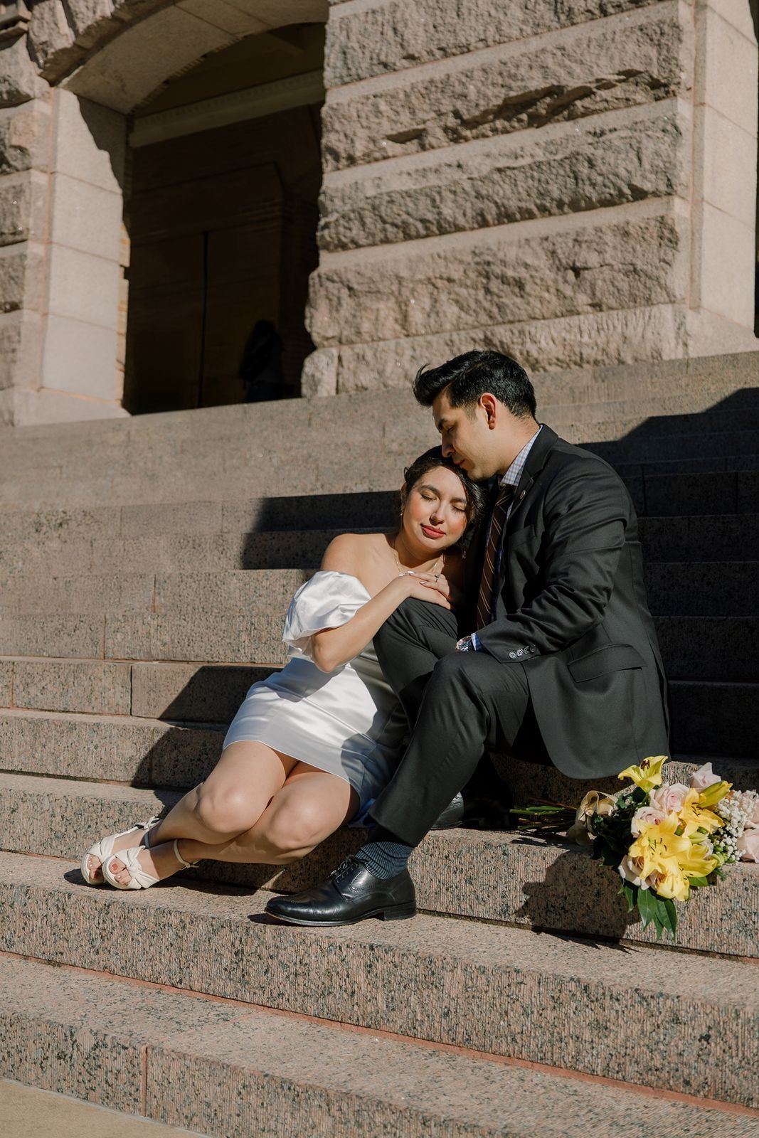 Bride and groom sit intimately on stone steps with a yellow bouquet beside them on their wedding day.