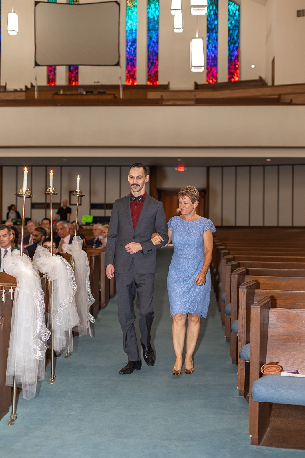 Smiling groom escorts woman in blue dress down church aisle at wedding.