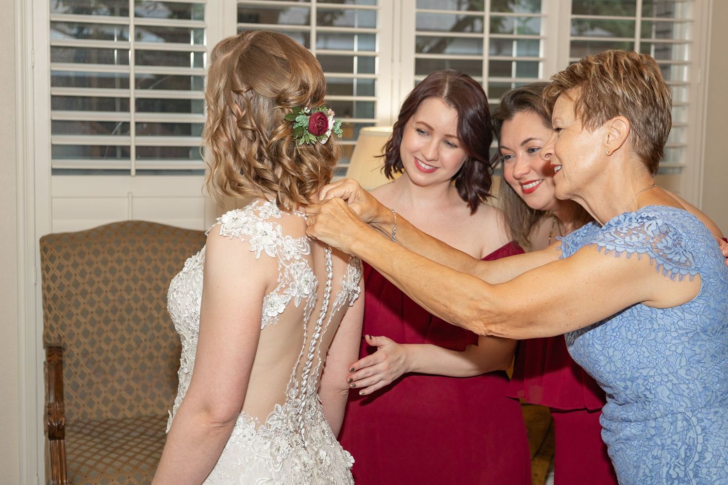 Bridesmaids and mother help bride with lace wedding dress buttons in preparation room.