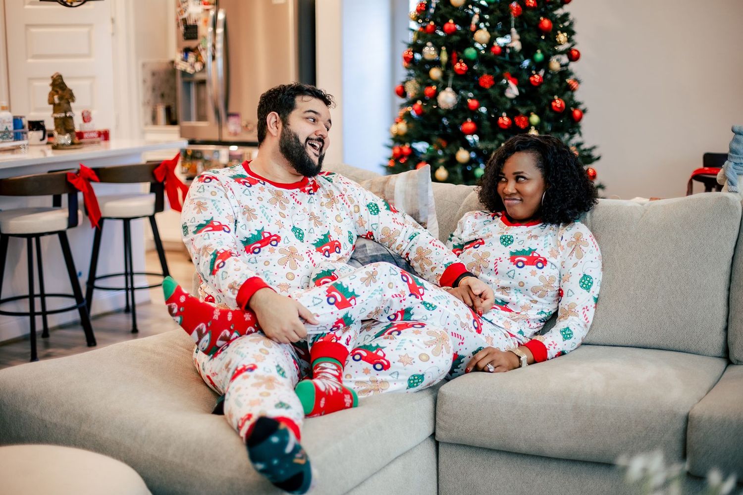 Couple in matching Christmas pajamas relaxing on gray couch with decorated tree in background.