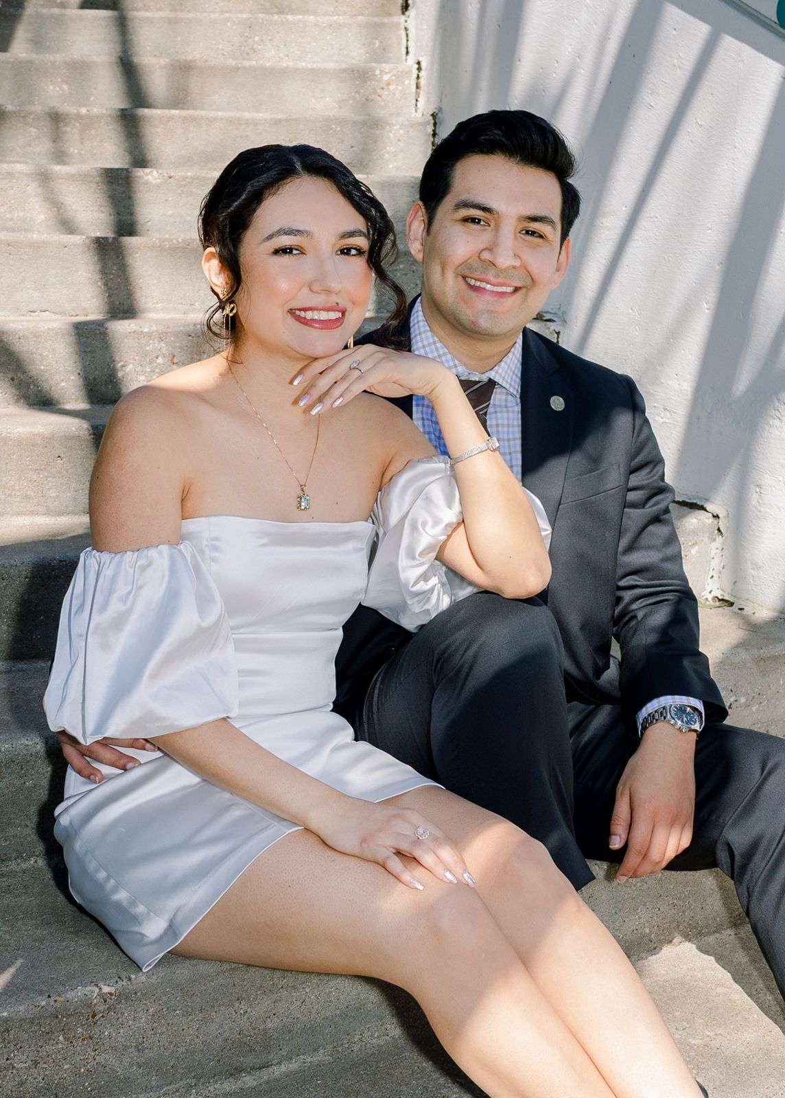 A couple in wedding attire sits together in an intimate photoshoot against a light-filled backdrop.