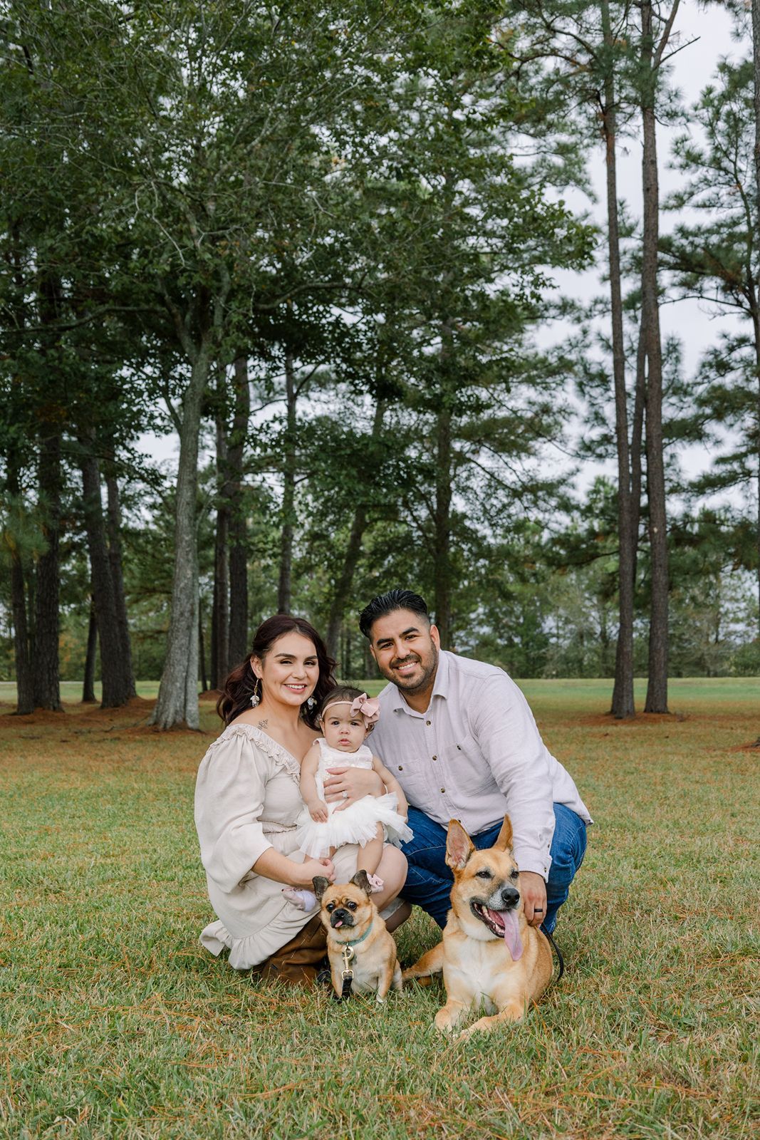 Family and their two dogs pose together on grassy lawn with tall pine trees in background.