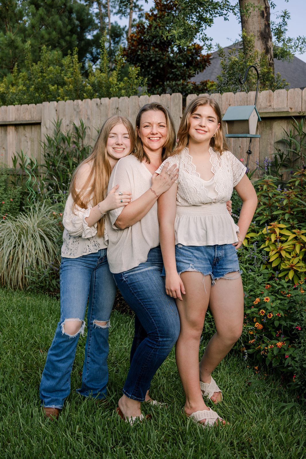 Three people stand together smiling in a backyard garden with a wooden fence and greenery in the background.