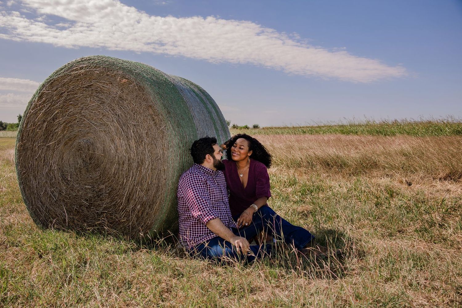 Couple sits together in golden field with hay bales during sunset creating romantic rural scene.