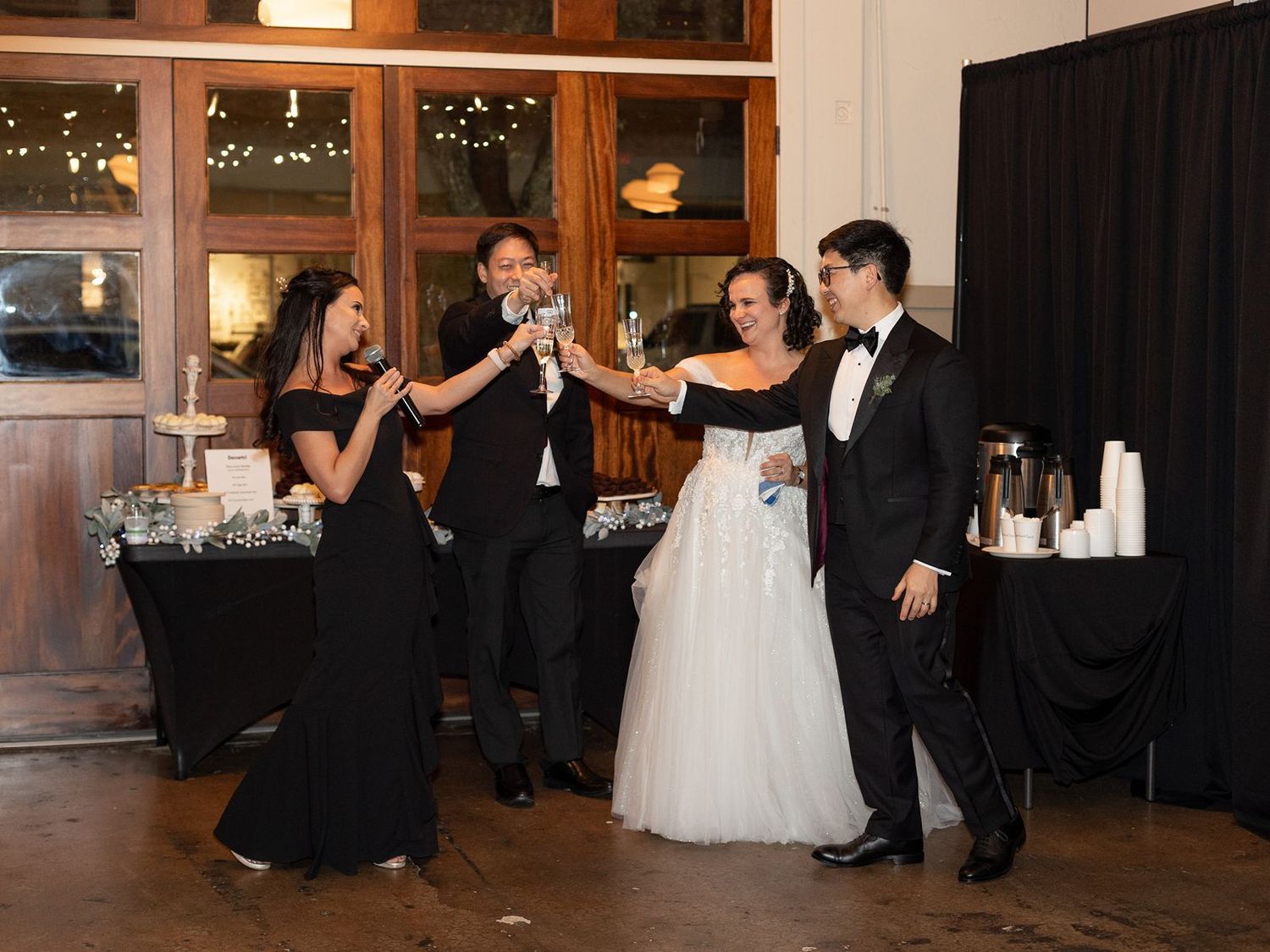 Wedding party celebrates with a joyful toast at an indoor reception venue with warm lighting and rustic decor.
