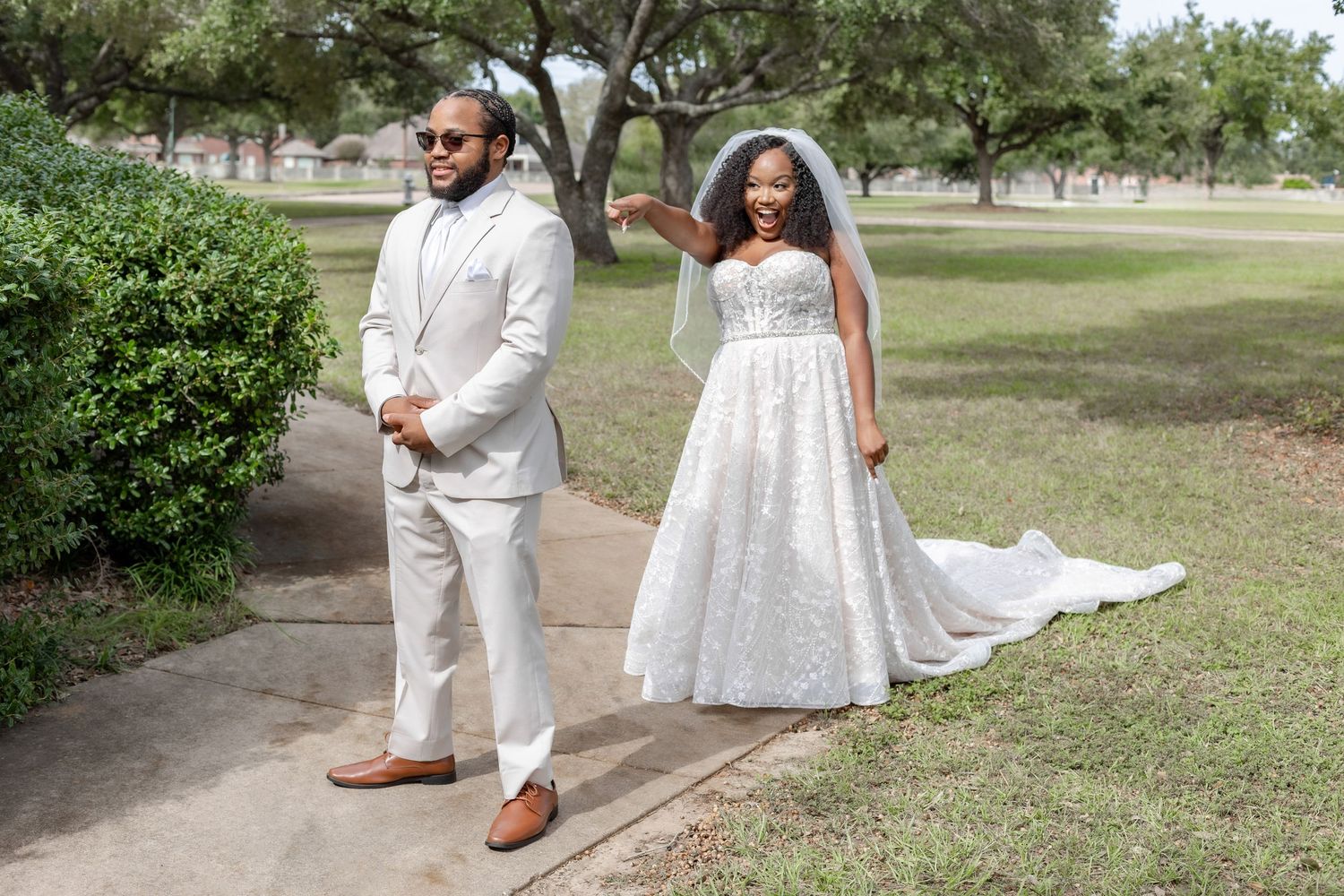 A couple in elegant white wedding attire poses together in a sunlit outdoor garden setting.