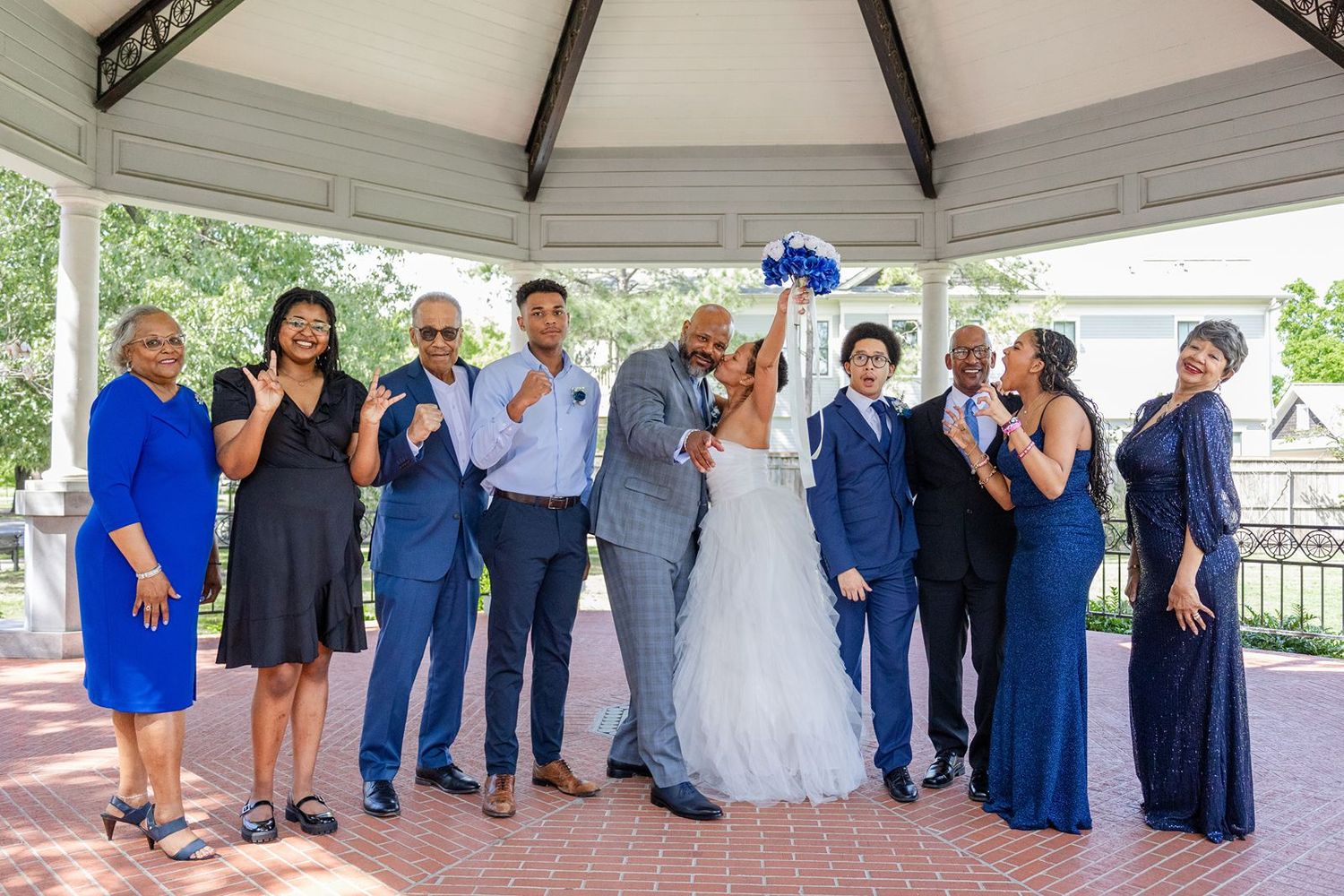 Wedding party poses under gazebo, bride and groom kissing while guests in royal blue attire celebrate with bouquet raised.