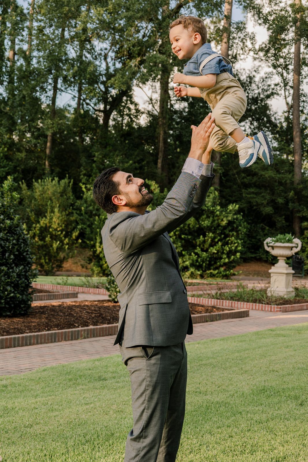 A man in a gray suit lifts a small child into the air on a manicured lawn with trees in the background.